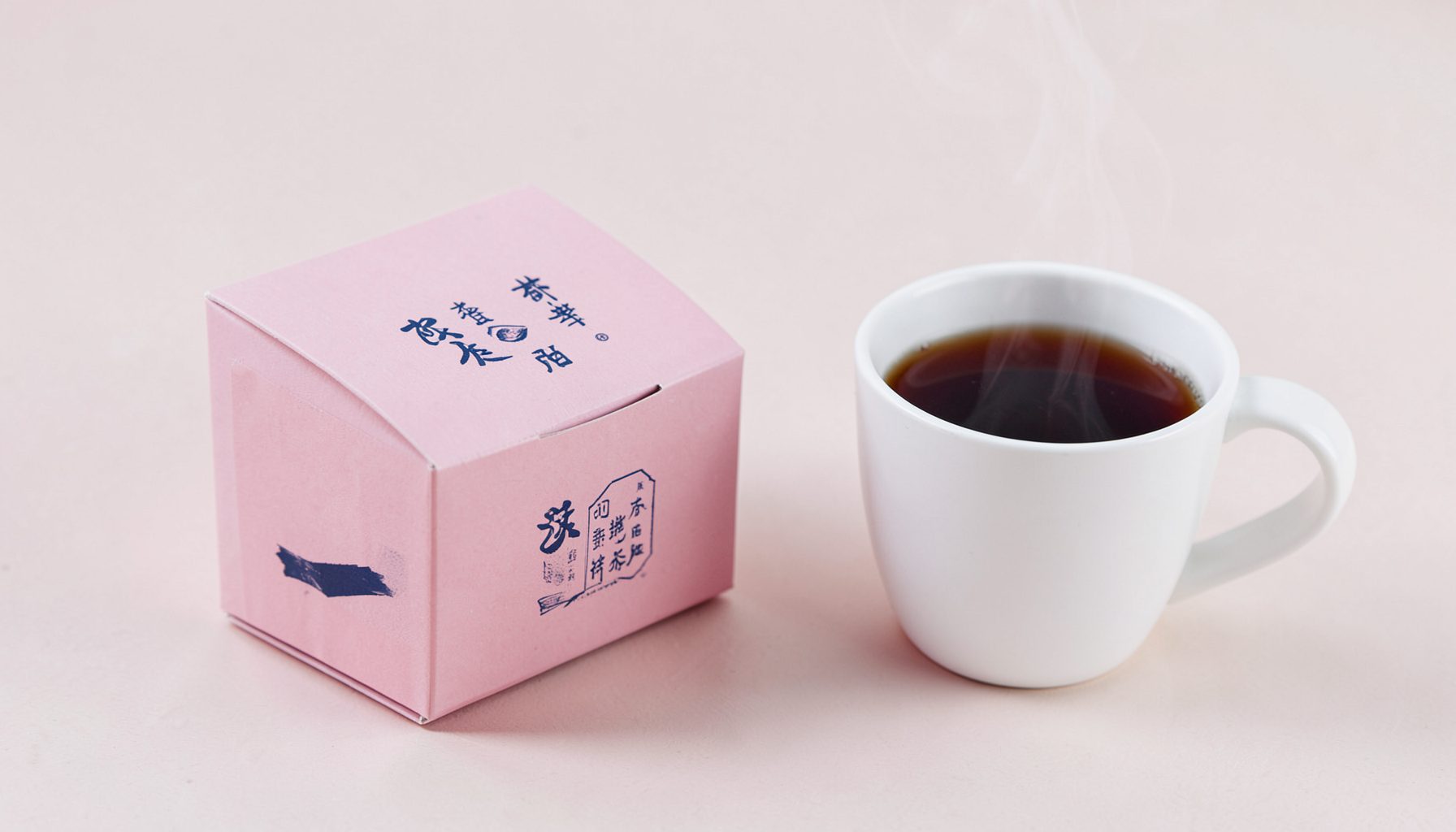 Small paper box of tea bags in soft pink and navy packaging beside a white cup of steaming dark tea